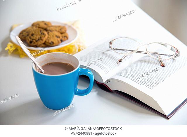 Cup of chocolate, open book, eyeglasses and biscuits