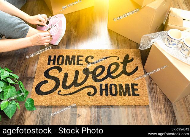 Woman wearing sweats and untying pink shoes near home sweet home welcome mat, moving boxes and plant