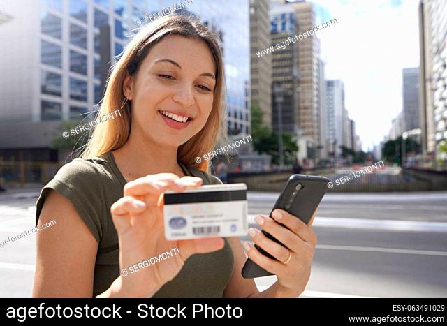 Beautiful young woman watching her credit card number using her smartphone for shopping online in finance district