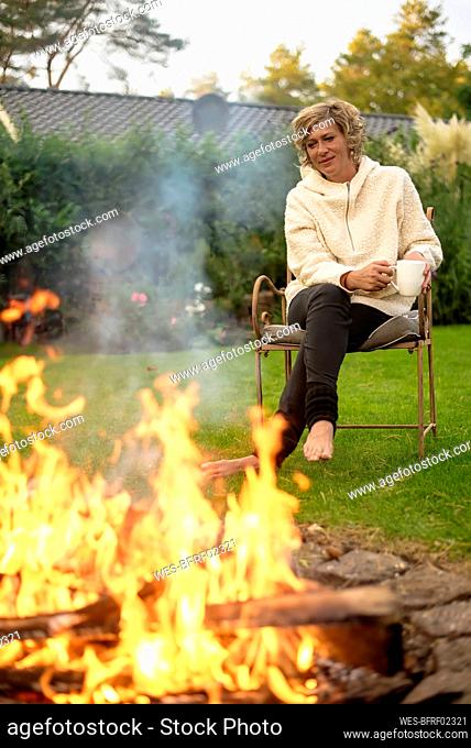 Mature woman with coffee cup looking at bonfire while sitting on chair at back yard