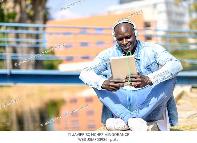 Man wearing casual denim clothes listening music with headphones and digital tablet outdoors