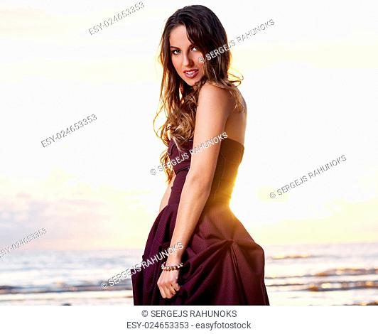Beautiful girl with brown hair who is wearing a dark purple dress is posing at a beach