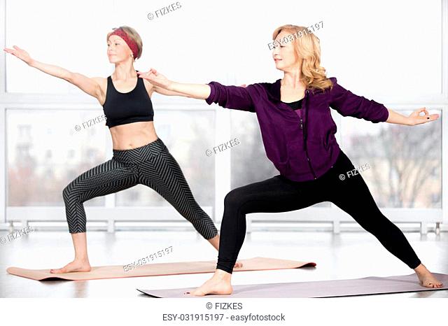 Fitness, stretching practice, group of two attractive happy smiling fit mature women working out in sports club, doing lunge, Warrior II posture