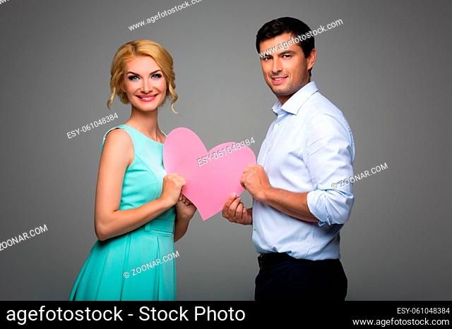 Beautiful young couple holding big pink hearts. Blond woman in mint dress and handsome man in shirt and trousers standing on grey background