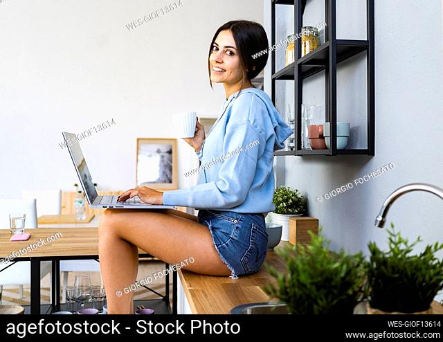 Smiling woman with laptop having coffee in kitchen at home