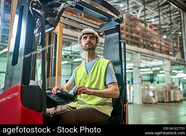 Serious concentrated man sitting at the steering wheel of the electric forklift and looking up