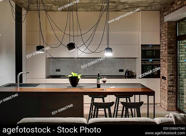 Kitchen in a loft style with concrete and brick walls. There is a kitchen island with a sink, plant and black chairs, light lockers with built-in oven and...