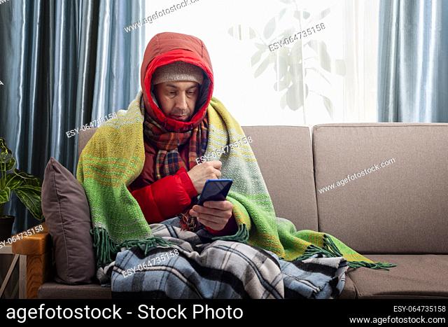Middle age male model in hat parka and plaid sitting on the sofa while using his smartphone in his cold living room