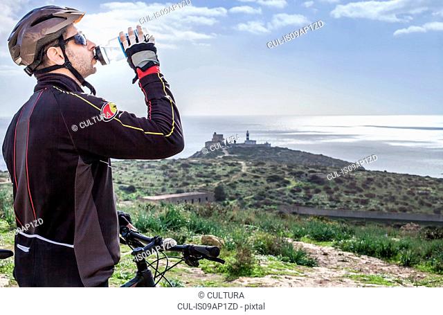 Mid adult male mountain biker drinking water on coastal path, Cagliari, Sardinia, Italy