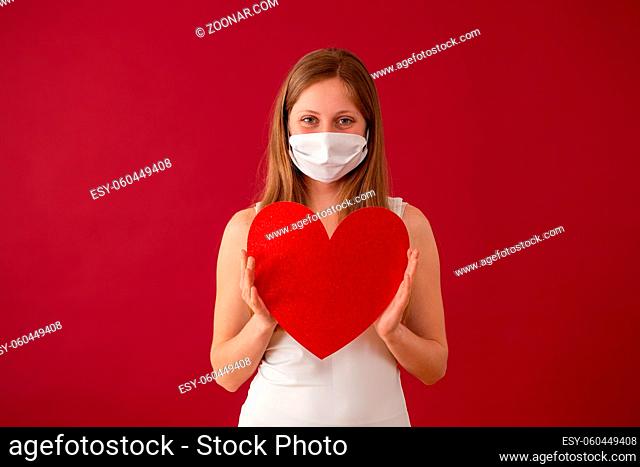 Smiling woman wearing face mask and holding red heart in hands. Concept of valentine during coronavirus pandemic. Cheerful blonde Caucasian girl from front view