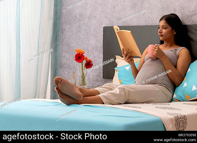 Pregnant woman drinking hot drink and reading a book while relaxing on bed at home