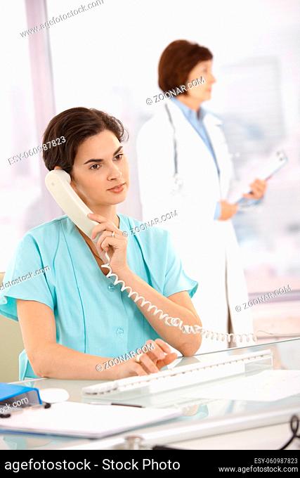 Assistant sitting at desk taking phone call using computer, medical doctor standing in background