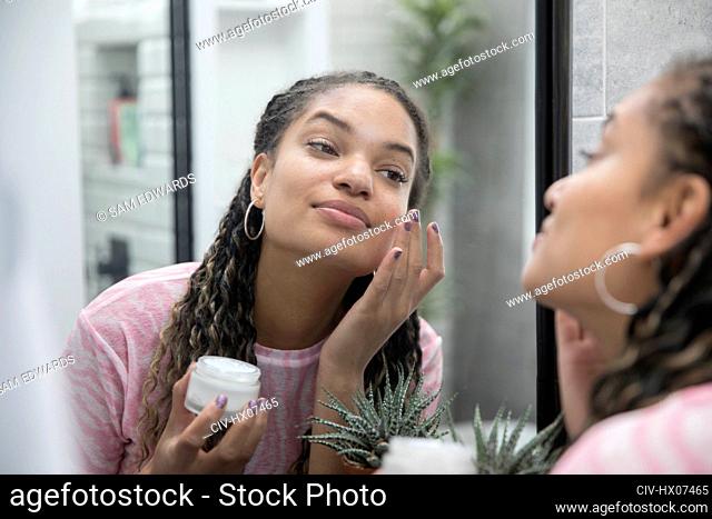 Beautiful young woman applying moisturizer in bathroom mirror