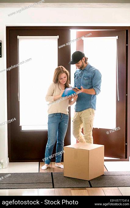 Female and male standing in hallway at home. Courier bring a cardboard box package. Customer signing receipt of the parcel