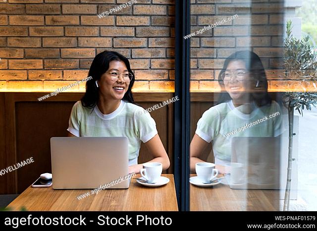 Young woman with laptop looking away while sitting near glass window at cafe