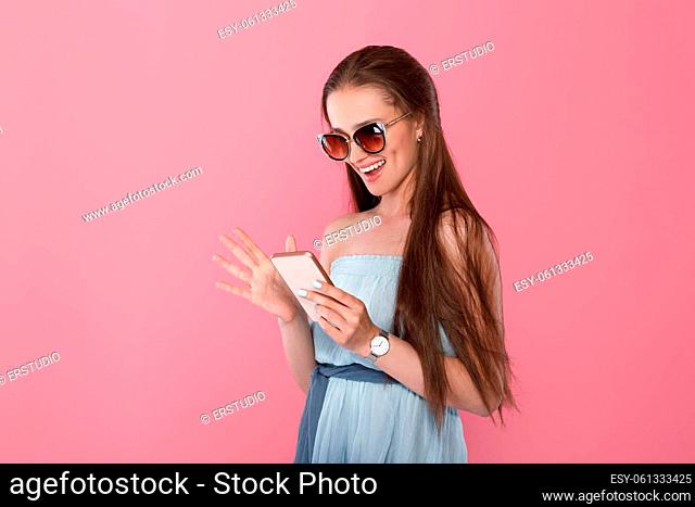 portrait of beautiful woman in sunglasses and blue dress using smartphone on pink background