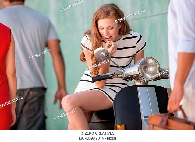 Young woman sitting on moped applying lipstick in city