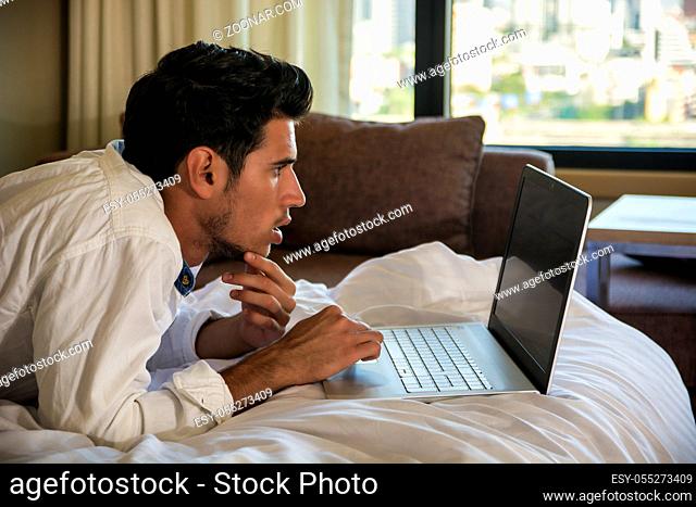 Attractive Young Man with Serious Expression, with Laptop on Bed Working on his Start-up Business - Young Male College or University Student Doing Homework
