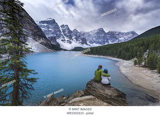 Asian couple sitting on rock admiring scenic view of mountain lake