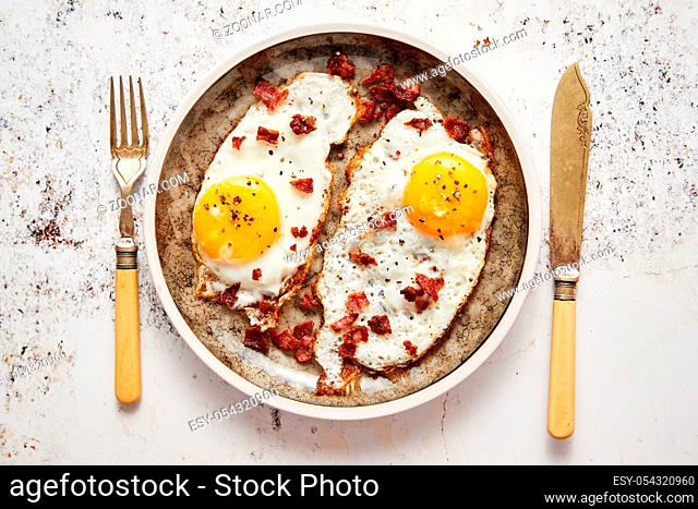 Two fresh fried eggs with crunchy crisp bacon served on rustic plate. Fork and knife on sides. Top view