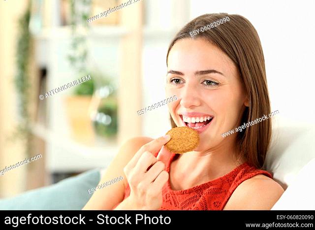 Happy woman is eating cereal cookie sitting on a couch in the living room at home