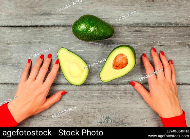 Tabletop view - woman hands with red nails, avocado cut in half, whole one above, on gray wood desk
