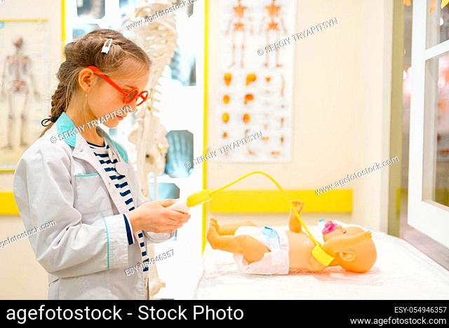 Little girl in uniform playing doctor with toy doll, playroom. Kid plays medicine worker in imaginary hospital, profession learning, childish dream