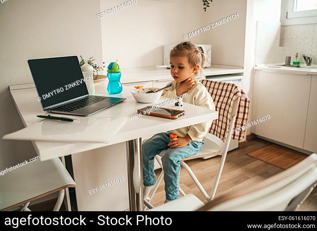 Serious pensive cute little Caucasian girl sitting alone at the table during the morning meal