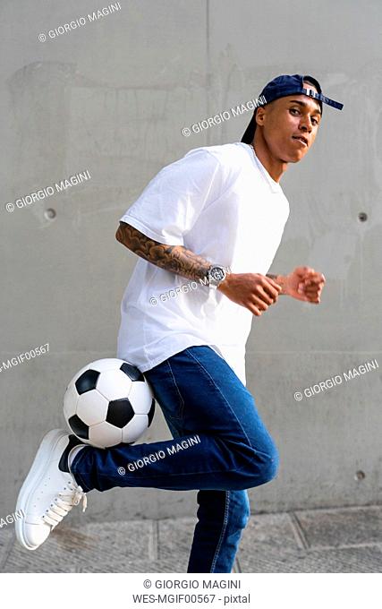Portrait of tattooed young man balancing football in front of concrete wall