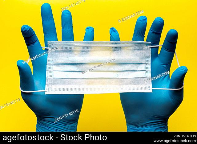 Doctor holds antivirus face mask in hands in blue medical gloves on yellow background. Pandemic insurance, airborne diseases, SARS, grippe