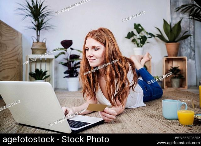 Woman with credit card doing online shopping through laptop while lying on floor at home