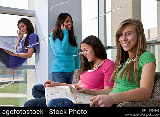 Four teenage girls in a high school corridor, one on a smart phone and others looking through textbooks