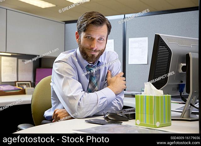 Portrait of young Caucasian man working in cubicle at office, fighting off a cold with tissues and hot tea, looking at camera smiling