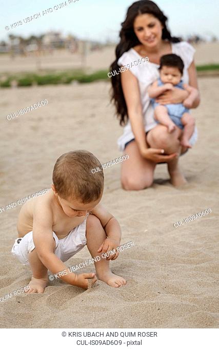 Toddler digging in sand with hand, mother and baby in background