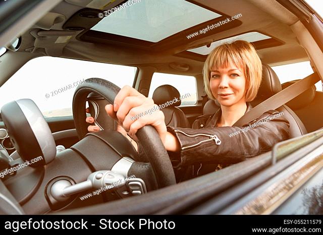 A cute young woman with a short haircut sits behind the wheel of a small car. The concept of driving a woman. Woman driving