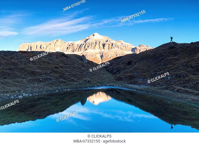 Hiker in silhouette near a small pond with Sella group with the highest Piz Boè mountain reflected, Pordoi pass, Arabba, Beuuno, Veneto, Italy