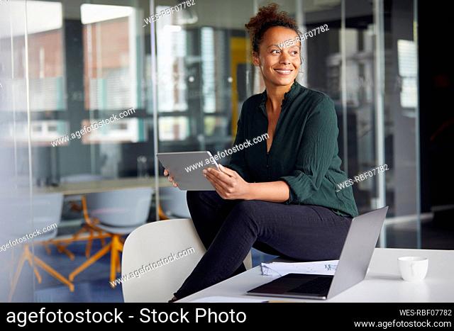 Portrait of smiling businesswoman with digital tablet sitting on desk looking at distance