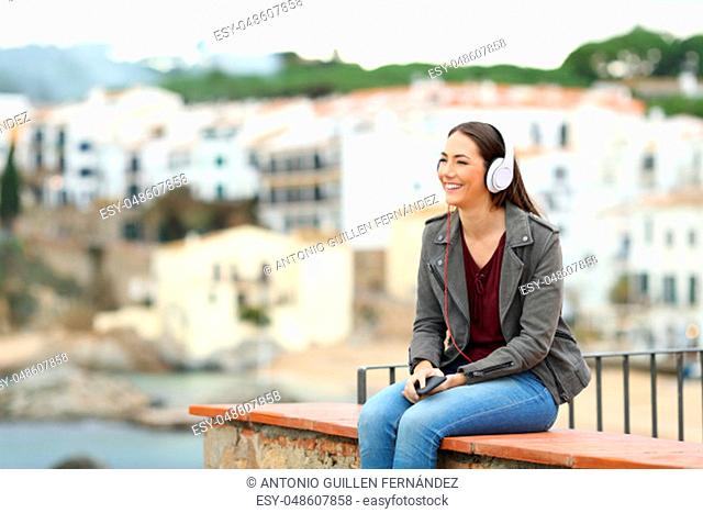 Happy woman relaxing listening to music wearing headphones sitting on a ledge on vacation in a coast town