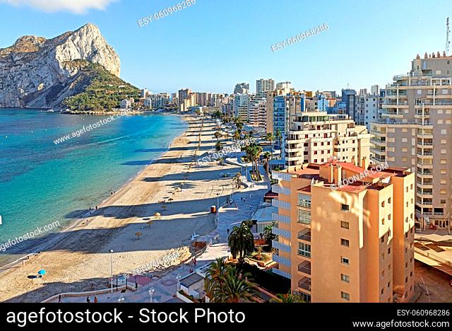 Sandy beach with parasols and vacationers, Penyal d'Ifac Natural Park view. Turquoise blue Sea water summer day. Seafront promenade of Calpe spanish resort town