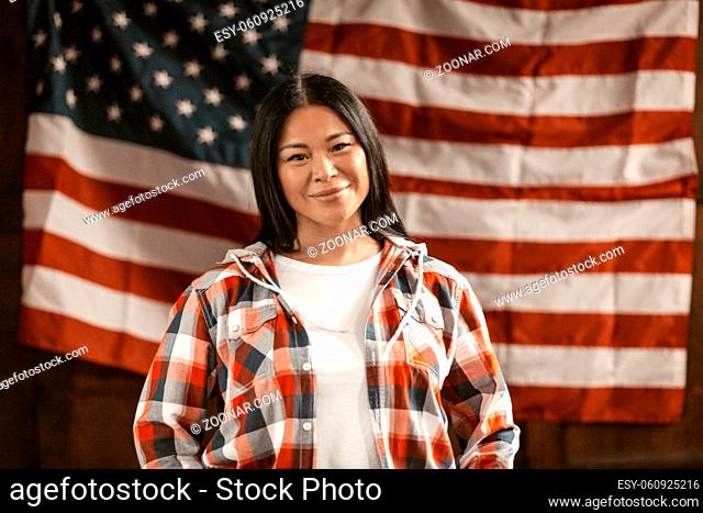 Cute Asian Woman Smiles On America's Flag Background, Smiling Brunette In Plaid Shirt Indoors Concept Of Patriotism Front View
