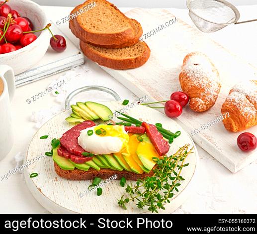toast with poached egg and avocado on a round board, next to croissants and ripe red cherries, morning breakfast, top view on a white table