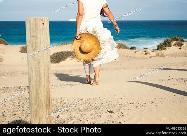 Elegant fashion lady in white dress and natural hat walking to the beach barefoot enjoying the summer holiday vacation day in tropical place - blue sea and sky...