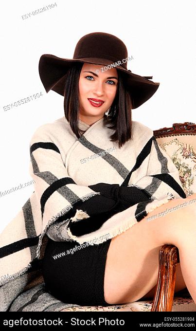 A lovely young woman sitting in an old armchair with her legs over the side, wearing a brown hat and poncho, isolated for white background