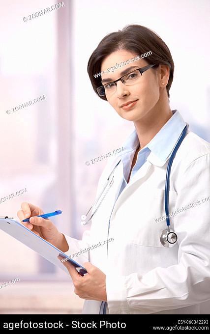 Attractive female doctor standing on hospital corridor doing paperwork, looking up at camera