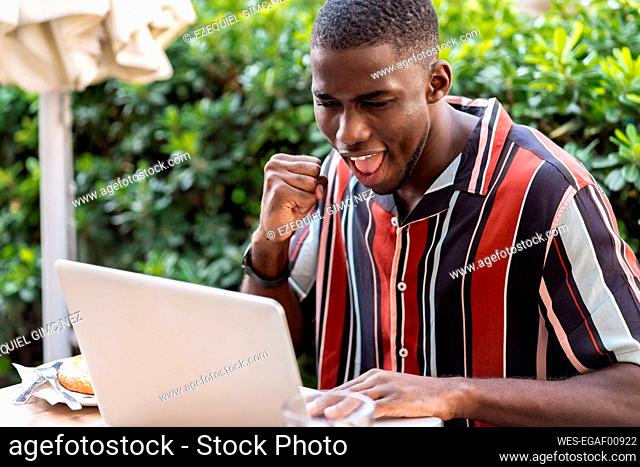 Happy young man clenching fist while using laptop on terrace of cafe