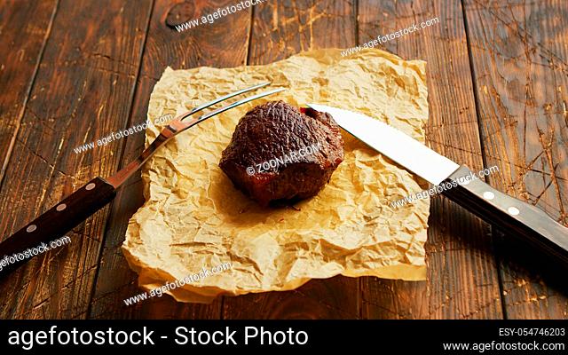 From above shot of fork and knife lying near parchment paper and piece of delectable fried meat on scratched lumber tabletop