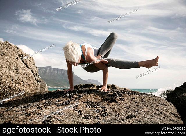 Blond woman practicing Astavakrasana on rock formation against sky