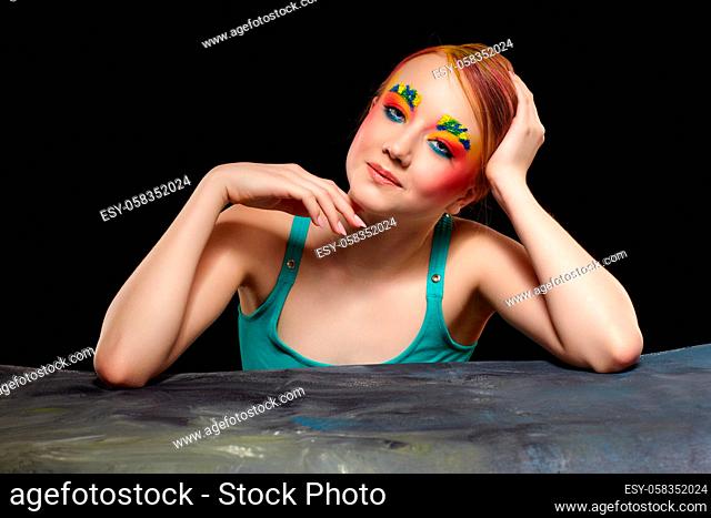 Teenager girl portrait with unusual face art make-up. Paint on brows and hair. Young female is sitting at a black table on black background