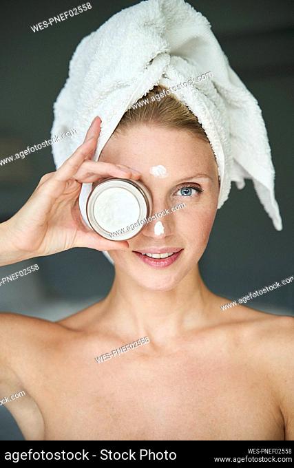 Portrait of beautiful woman with head wrapped in a towel holding cream jar in front of her eye