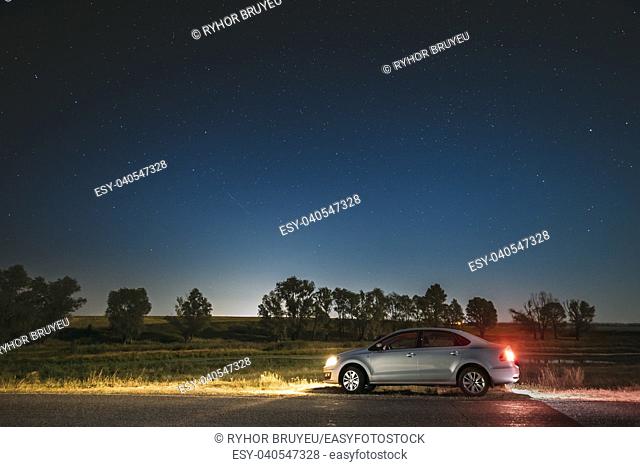 Blue Night Starry Sky Above Country Asphalt Road In Countryside. Sedan Car Parking Near Asphalt Road In Summer Night. Yellow Car Lights And Night View Of...